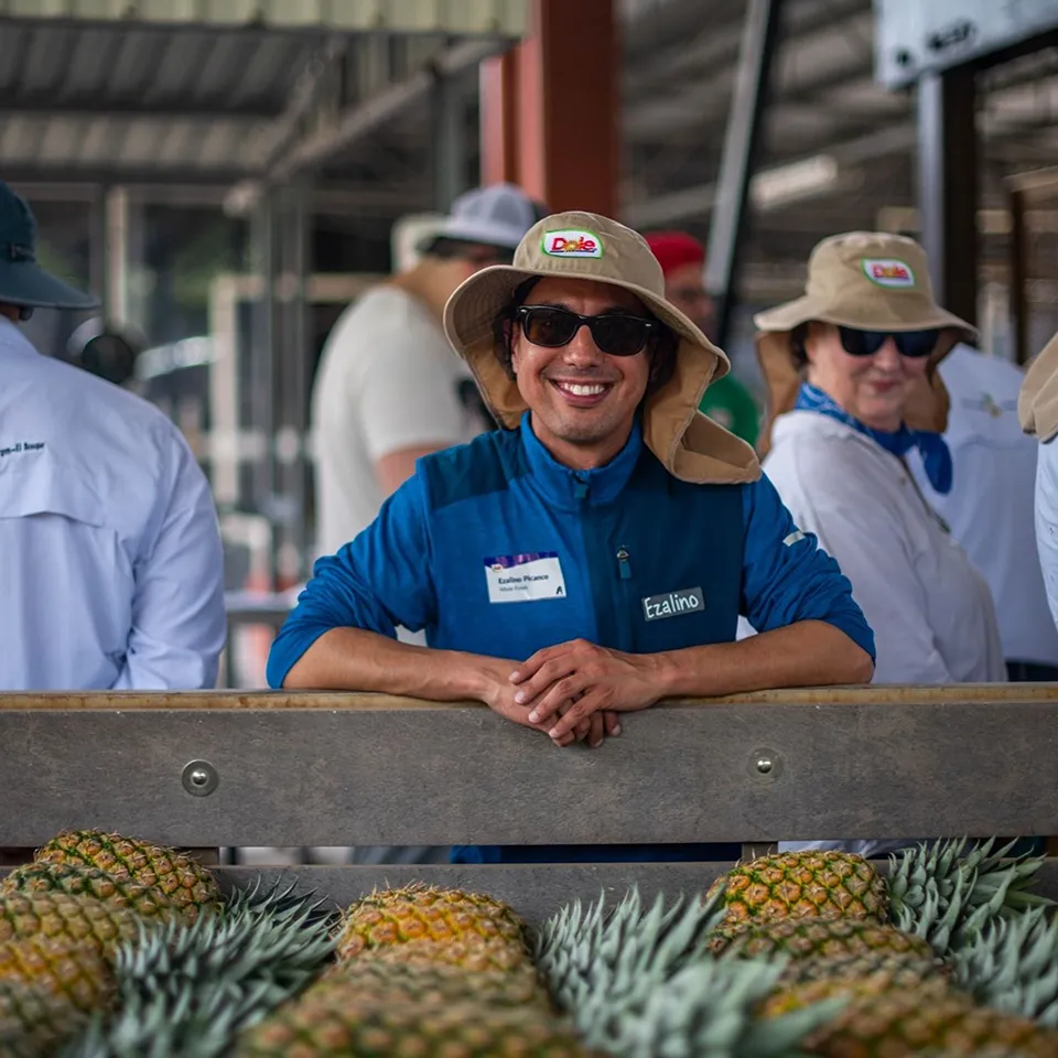 Ezalino, Store Trainer, smiling in a hat and sunglasses leaning on a surface with pineapples.