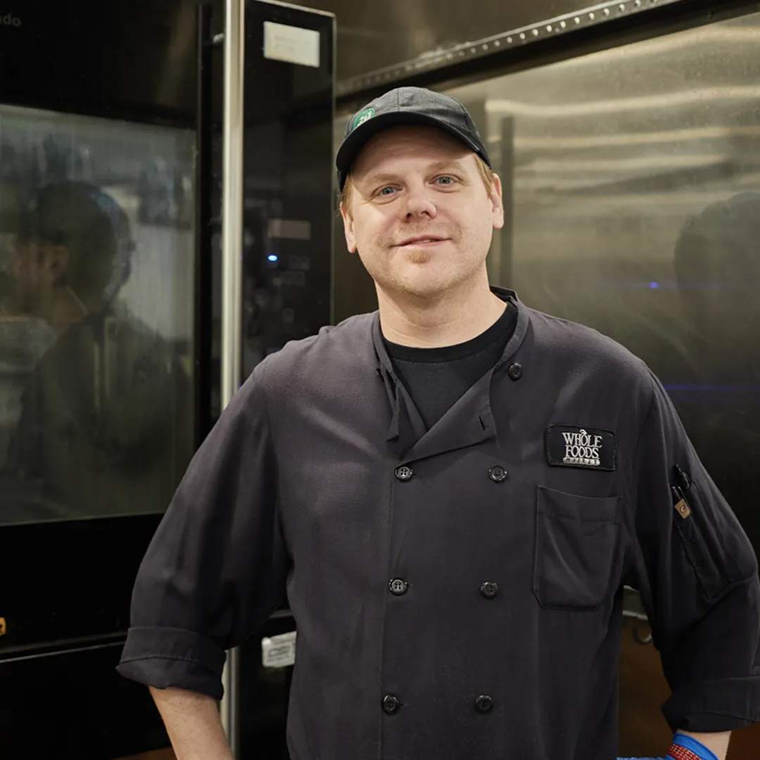 Whole Foods chef in black uniform smiling in front of ovens