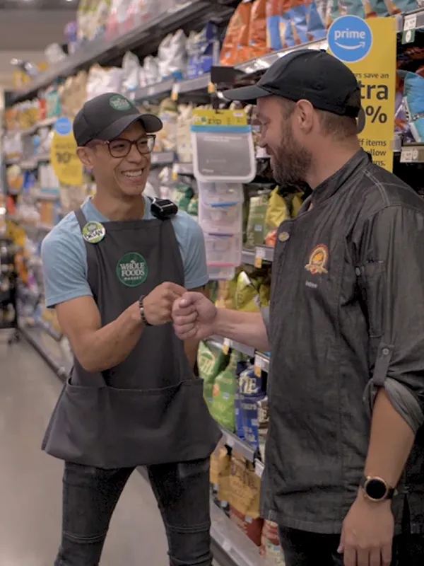 Two Whole Foods employees bumping fists in a grocery store, possibly promoting wellness programs.