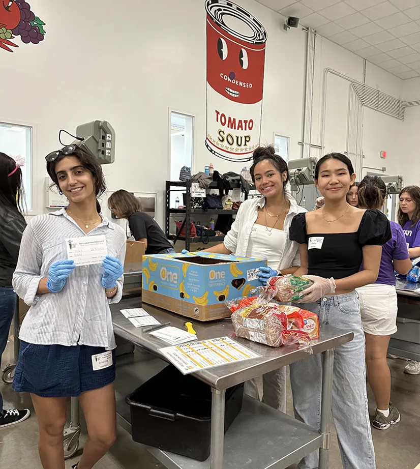 Three young women preparing food donations at a community event, a check and donations are visible.