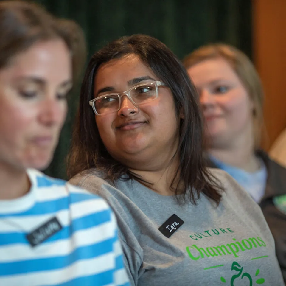 Iqra, wearing glasses, smiling, and wearing a gray t-shirt with the words Culture Champions.