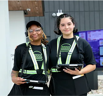 Two Shake Shack employees wearing uniforms and holding tablets stand together outside a Shake Shack restaurant