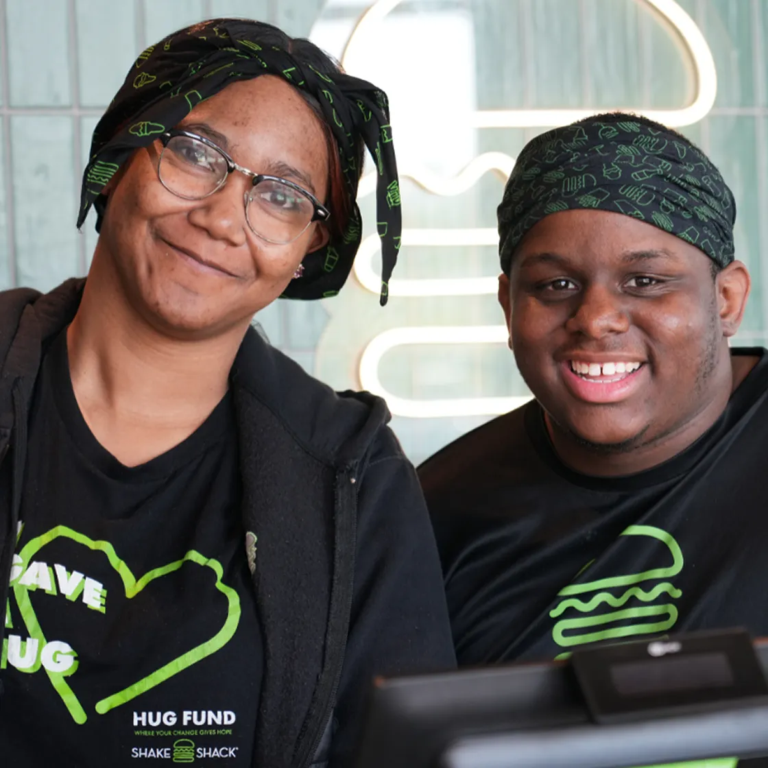Two Shake Shack employees smiling, wearing black shirts with the Hug Fund logo