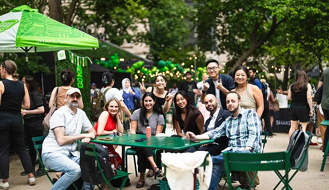 Happy diverse group of people sitting at a table outdoors under a green umbrella at a Shake Shack support center event
