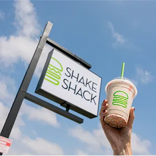 A hand holds a Shake Shack milkshake in front of a Shake Shack sign