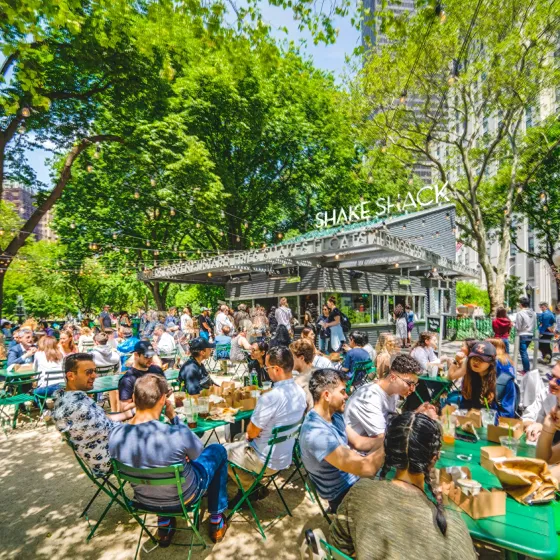 Crowded outdoor Shake Shack with people seated at green tables under trees