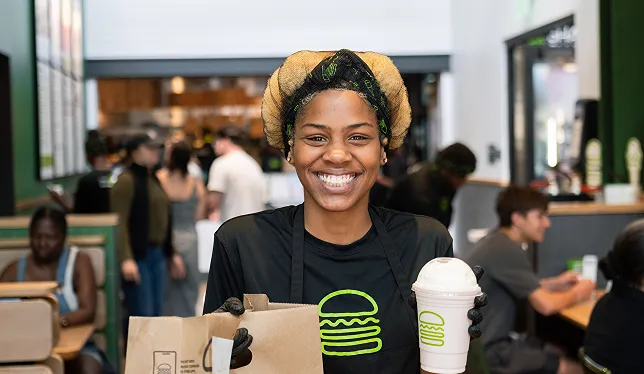 A smiling Shake Shack employee holds a Shake Shack order bag and a milkshake