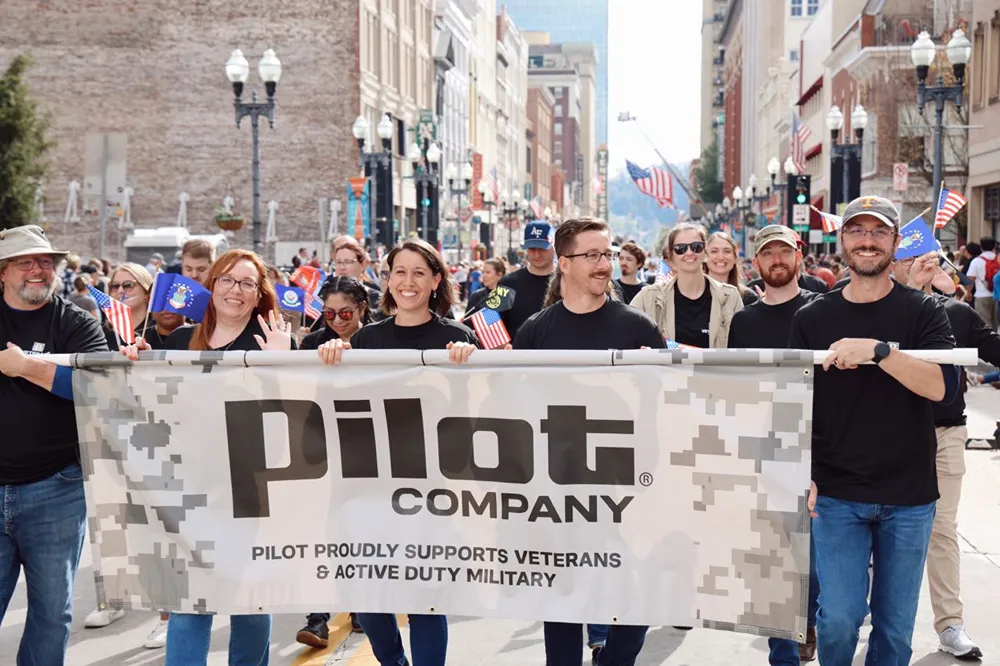 A group of smiling Pilot Company employees is marching in a parade with a banner supporting military veterans.