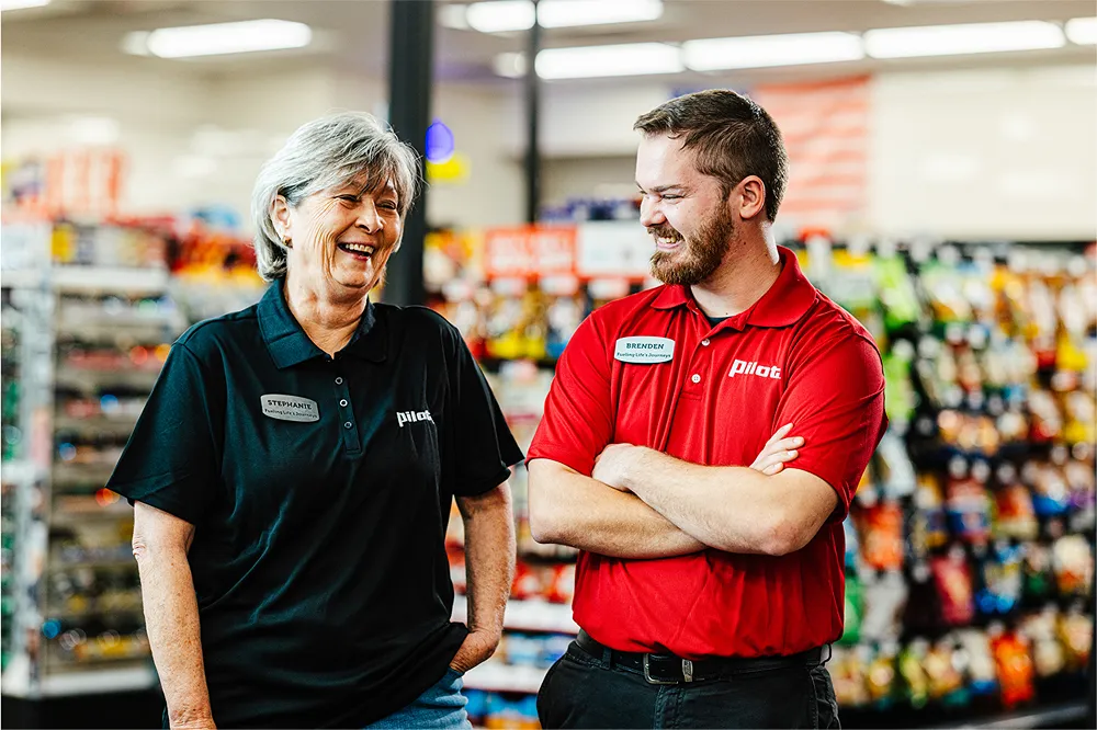 Two cheerful Pilot Flying J team members, a man and a woman, are smiling and chatting in a brightly lit store aisle.
