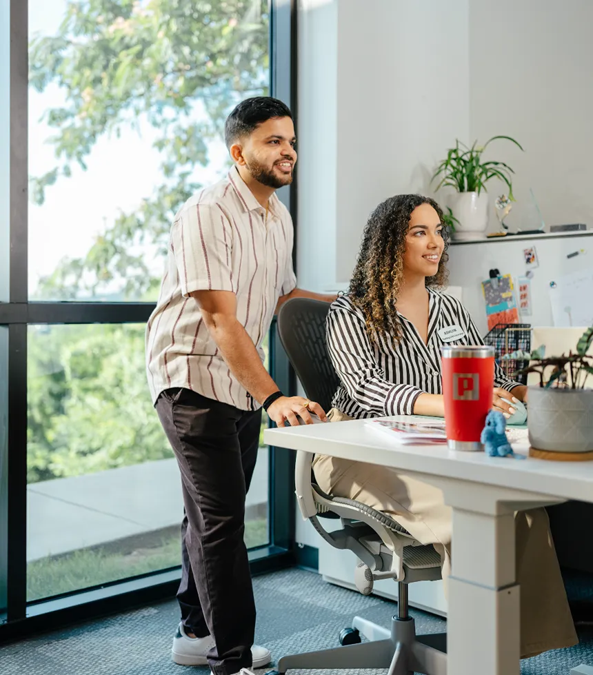 Two corporate employees are collaborating at a desk with multiple computer screens in a large, busy office.