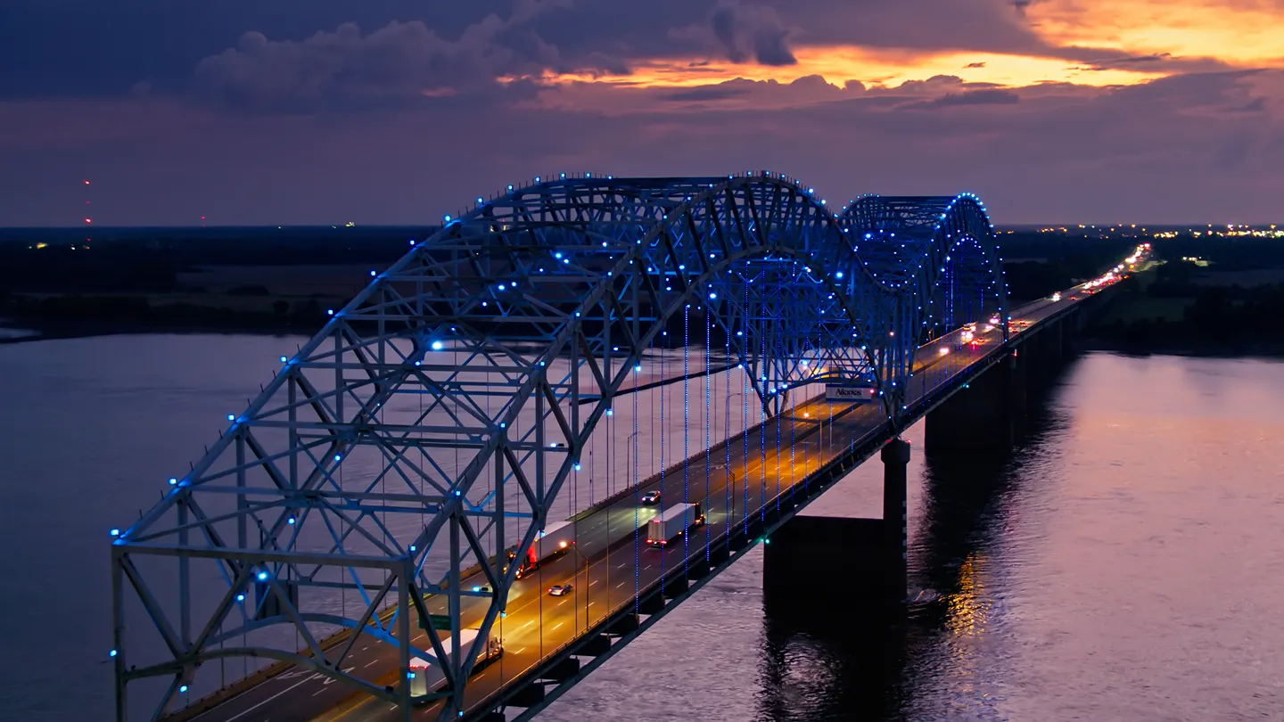 An aerial view of a large steel arch bridge illuminated with blue lights at sunset.