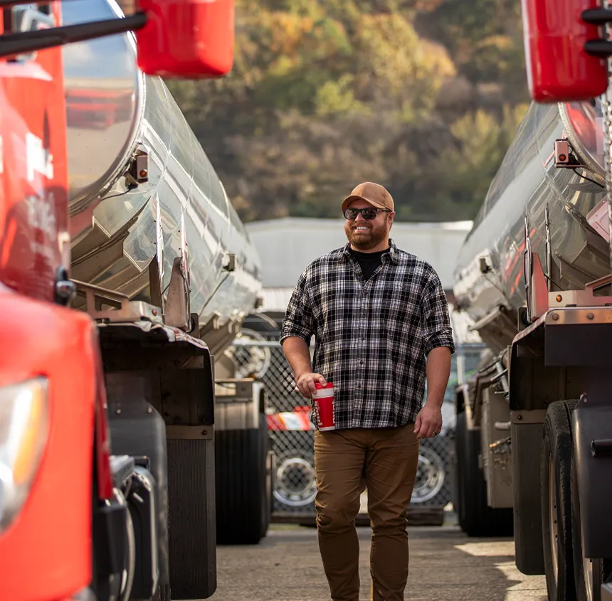 A smiling truck driver is walking between two fuel tanker trucks holding a coffee cup.