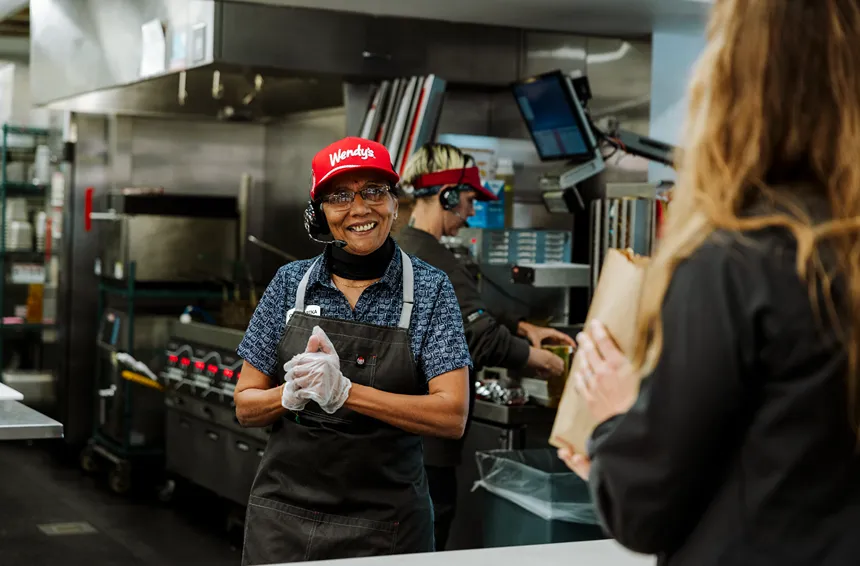 A smiling Wendy's fast-food employee is handing an order bag to a customer at the counter.