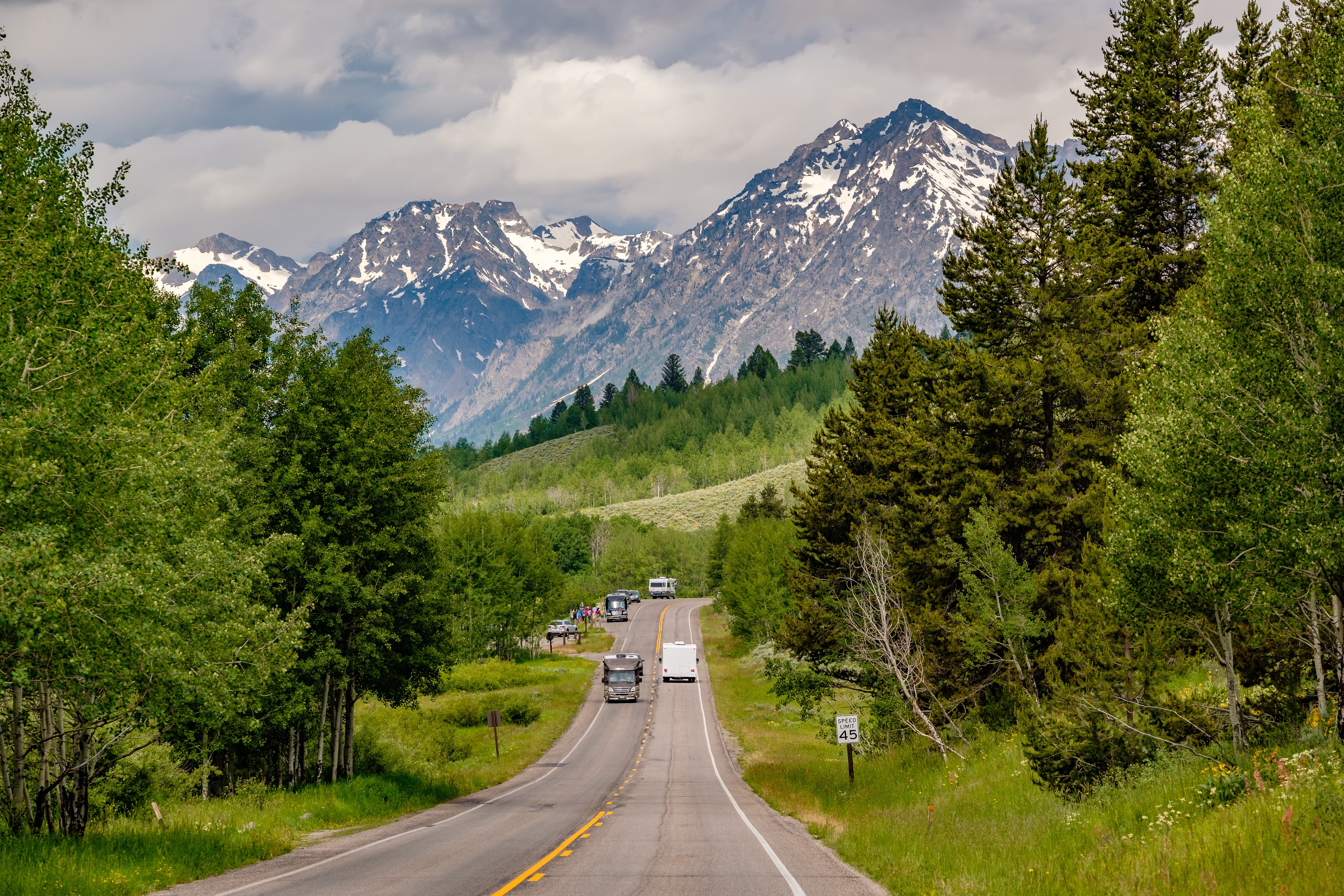 An inspiring view of a road leading toward snow-capped mountains through a lush green forest with several RVs traveling on it.