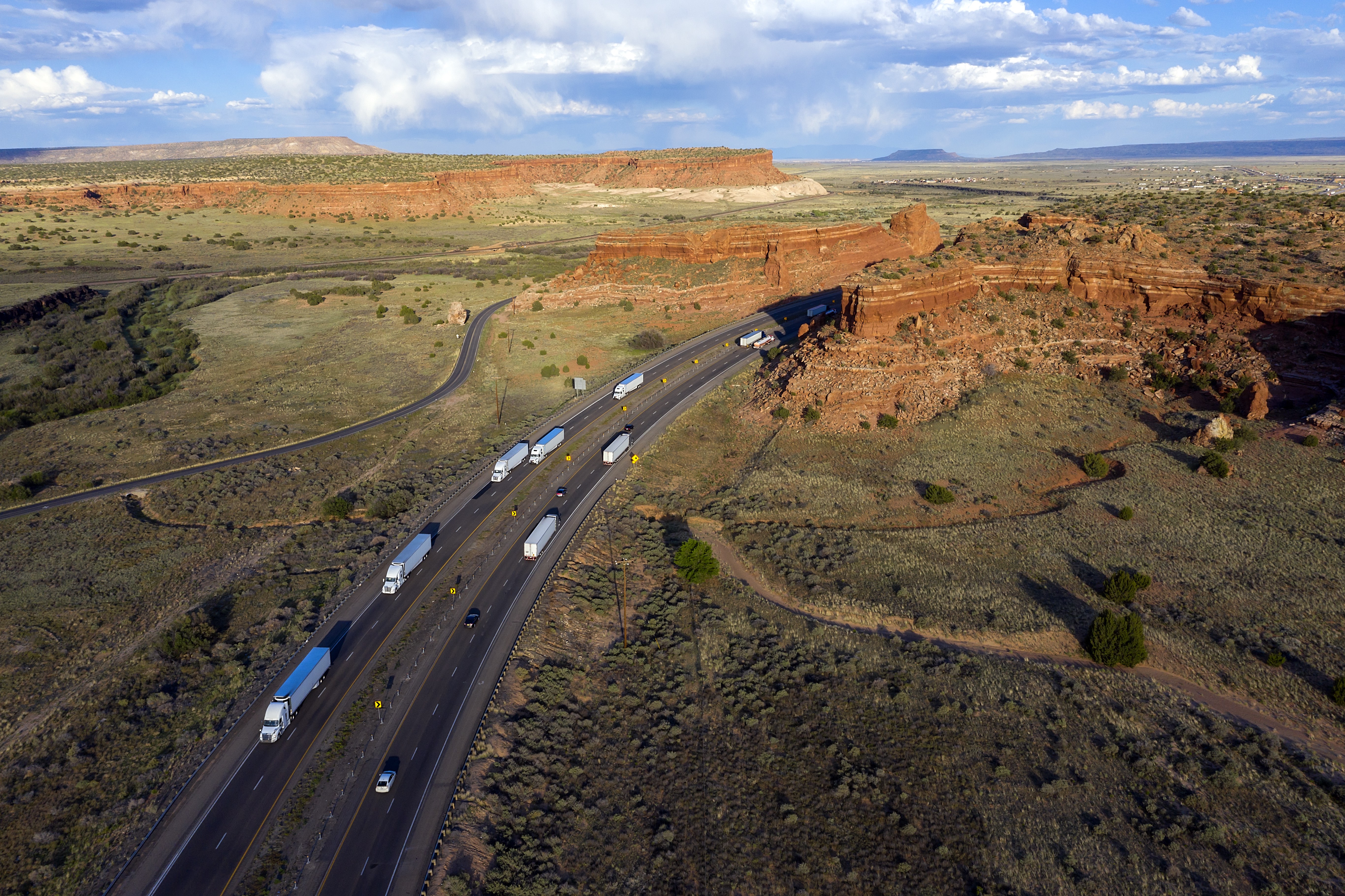 An aerial view of a highway with semi-trucks traveling through a vast red rock desert landscape.
