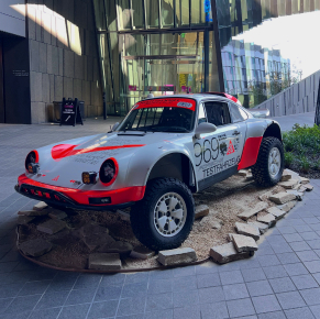 一輛銀紅色相間的保時捷911越野拉力賽車，編號969，配備升高懸吊和大型越野輪胎，被展示在現代建築內模擬的岩石地形底座上。