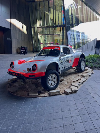 一輛銀紅色相間的保時捷911越野拉力賽車，編號969，配備升高懸吊和大型越野輪胎，被展示在現代建築內模擬的岩石地形底座上。