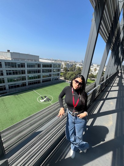 一位戴著太陽眼鏡的女子在戶外走道擺姿勢拍照，俯瞰下方綠意盎然的運動場。