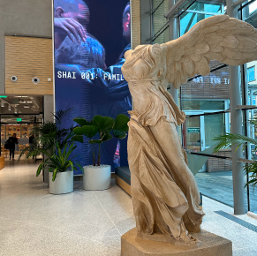 一座大型古典風格的薩莫色雷斯勝利女神（尼刻）雕像矗立在現代建築的大廳裡，大廳擁有巨大的窗戶，背景中還有一塊數位螢幕。