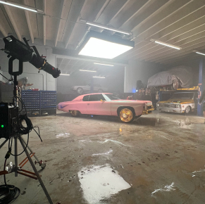 鏡頭切換到拍攝現場，展現了一間光線昏暗的車庫，裡面停著一輛經典的粉紅色低底盤車，配有金色輪圈。