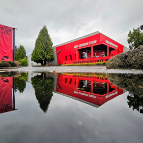 低角度拍攝的紅色建築和高大的常綠樹，完美地倒映在前景地面上的一個大水坑中。