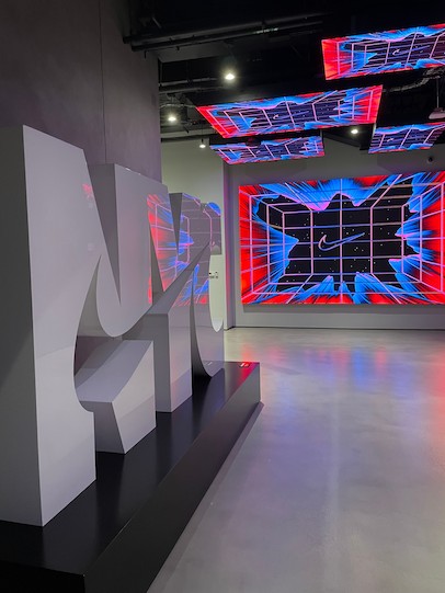 在一個昏暗的現代展廳裡，矗立著一座大型白色3D標誌雕塑，牆壁和天花板上的大螢幕投射出鮮豔的紅藍抽象網格圖案，為其提供照明。