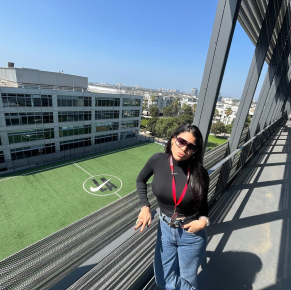 一位戴著太陽眼鏡的女子在戶外走道擺姿勢拍照，俯瞰下方綠意盎然的運動場。