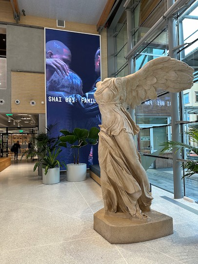 一座大型古典風格的薩莫色雷斯勝利女神（尼刻）雕像矗立在現代建築的大廳裡，大廳擁有巨大的窗戶，背景中還有一塊數位螢幕。