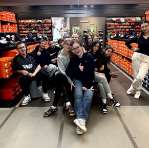 一群年輕人站在耐吉商店的走道上拍照，周圍的貨架上堆滿了橘色的鞋盒。