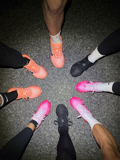 從上方俯視鏡頭，可以看到幾個人的腳圍成一個圓圈站在深色的人行道上，他們都穿著不同款式的耐吉跑鞋，顏色有亮粉色、橙色和黑色。