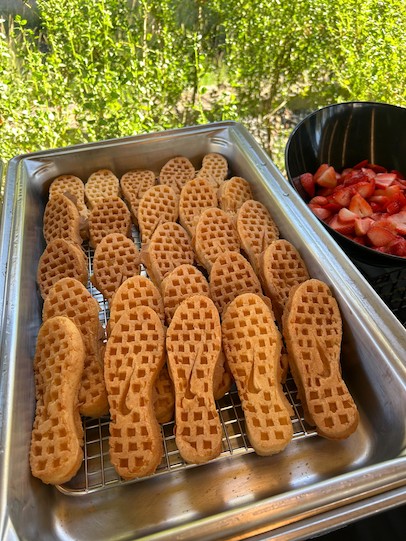 一個金屬烤盤，裡面裝滿了形狀像耐吉華夫格運動鞋鞋底的餅乾，放在金屬架上，旁邊是一個裝有切好草莓的黑色碗。