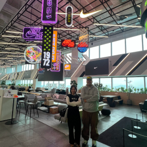 一男一女站在現代開放式辦公休息室裡，高高的裸露桁架天花板上懸掛著各種色彩鮮豔、霓虹閃爍的耐吉主題霓虹燈招牌。