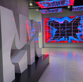 在一個昏暗的現代展廳裡，矗立著一座大型白色3D標誌雕塑，牆壁和天花板上的大螢幕投射出鮮豔的紅藍抽象網格圖案，為其提供照明。