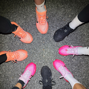 從上方俯視鏡頭，可以看到幾個人的腳圍成一個圓圈站在深色的人行道上，他們都穿著不同款式的耐吉跑鞋，顏色有亮粉色、橙色和黑色。