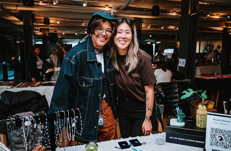Two people smiling behind a table with handmade jewelry at an indoor market