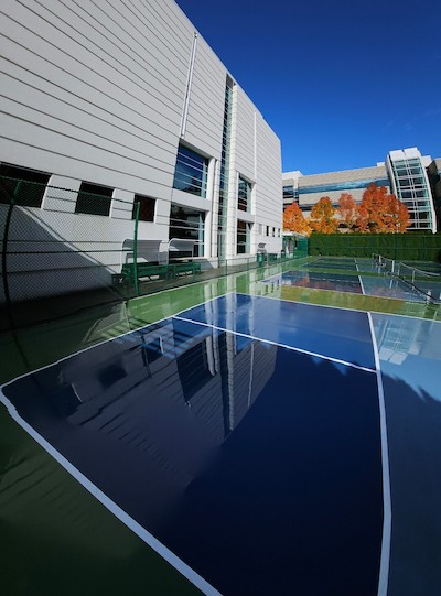 A reflective blue tennis court next to a modern building with autumn trees in the background