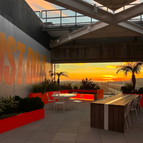Espace salon extérieur au coucher du soleil avec mobilier moderne, jardinières colorées et vue sur le coucher du soleil à travers une fenêtre en verre