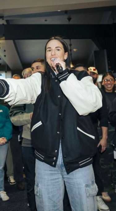 Een vrouw in een zwart-witte varsity-jas spreekt in een microfoon en gebaart naar een man die tegenover haar staat.