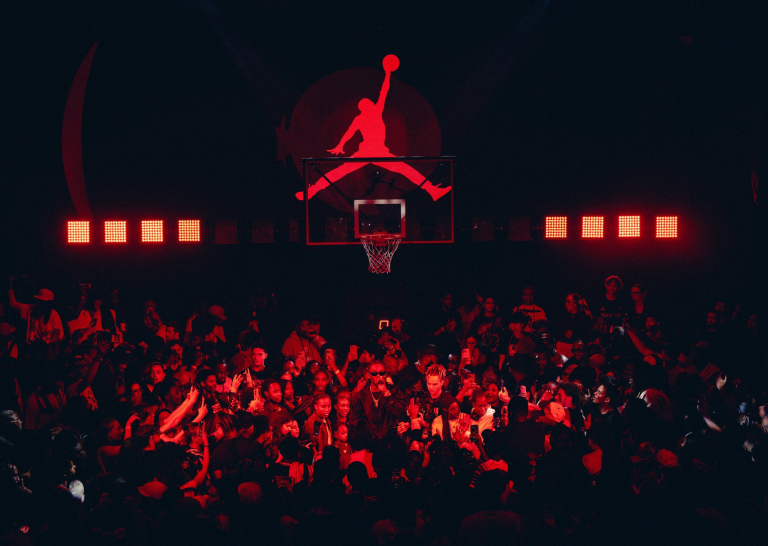 En fullsatt evenemangslokal med svag belysning badad i rött ljus, med en basketkorg i förgrunden och en stor, glödande röd Jumpman-logotyp projicerad på väggen bakom den.