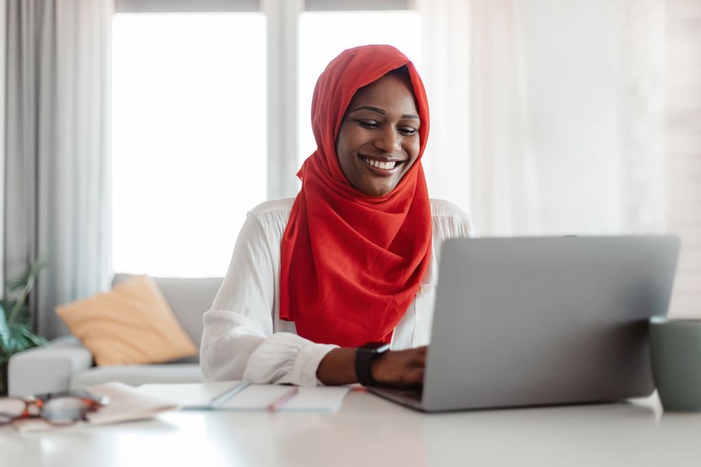 Person in a red hijab smiling at a laptop in a bright living room