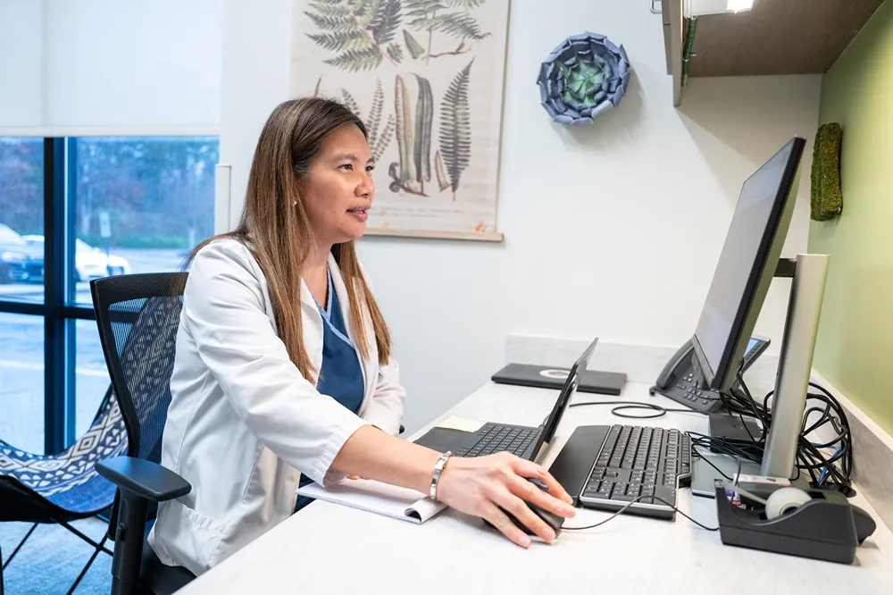 A woman in a white coat sits at a desk using a computer in a medical office. She appears focused, with medical charts and plants visible on the wall behind her.
