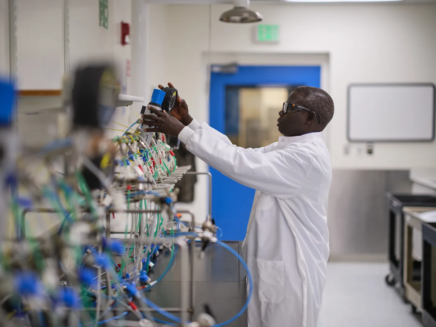 A scientist in a white lab coat adjusts equipment connected to multiple tubes and wires in a spacious laboratory with white walls and a blue door.
