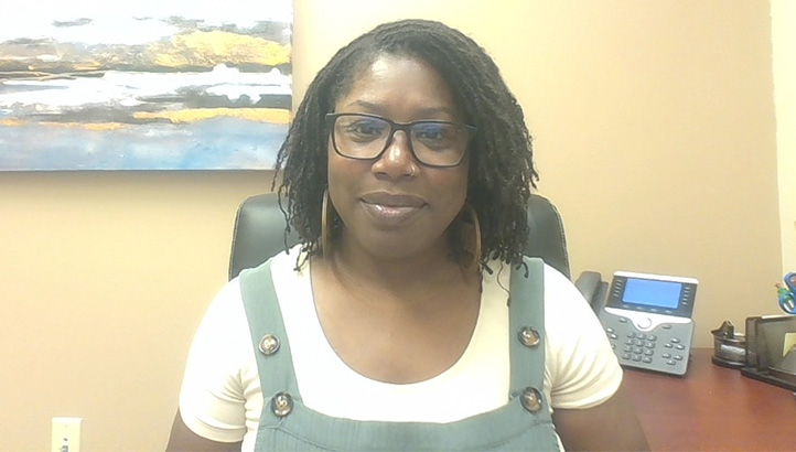 A woman with shoulder-length curly hair and glasses sits indoors, smiling slightly. She wears a cream shirt under green overalls. There is a painting with blue, white, and yellow colors on the beige wall behind her.