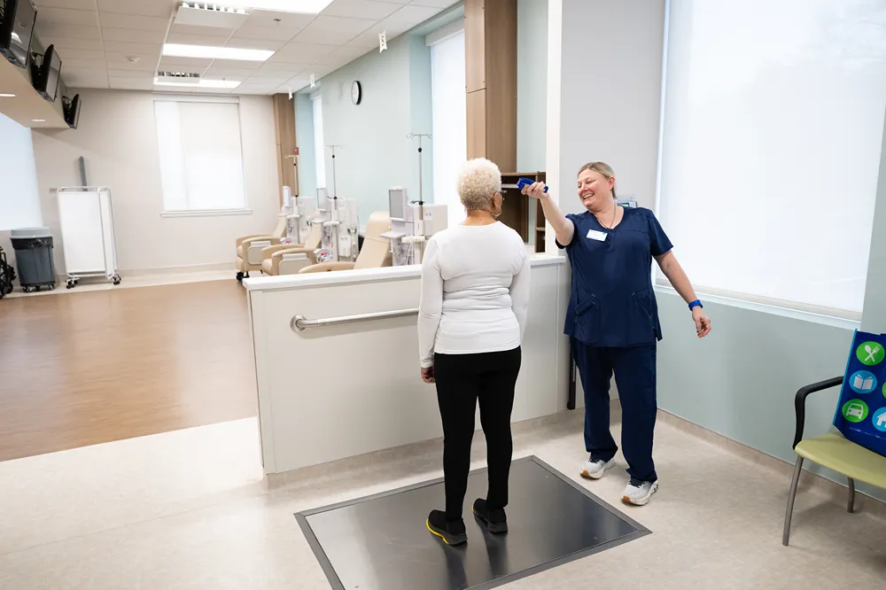 A healthcare worker in blue scrubs smiles while taking the temperature of an older woman with short white hair in a medical clinic with beige chairs in the background.