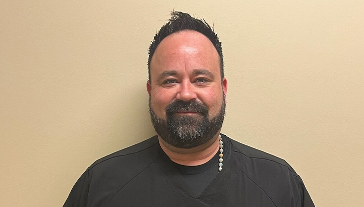A man with short dark hair and a beard is smiling, wearing a black shirt and standing against a plain beige wall.
