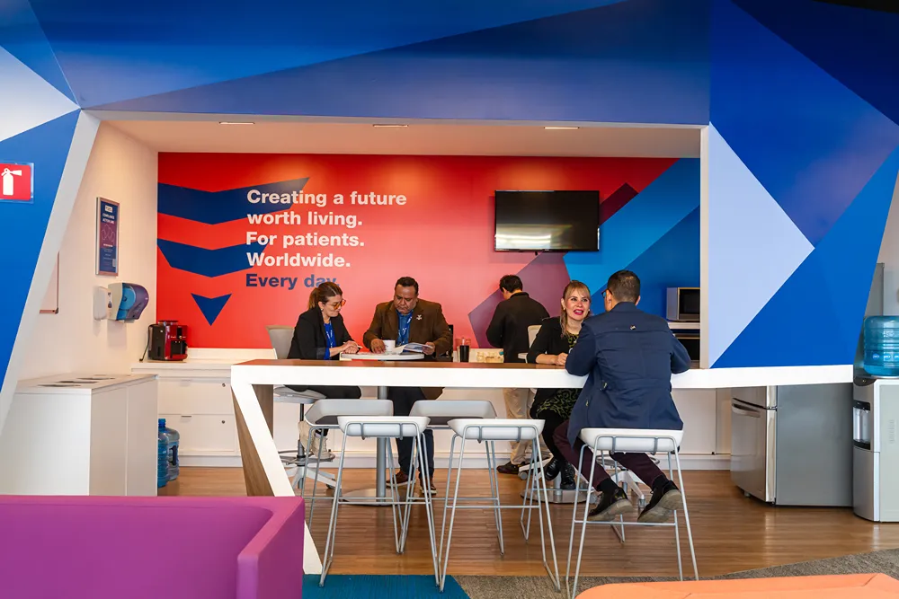 Five people sit and stand around a high table in a modern office lounge with colorful walls and a motivational quote about creating a better future for patients displayed on the back wall.