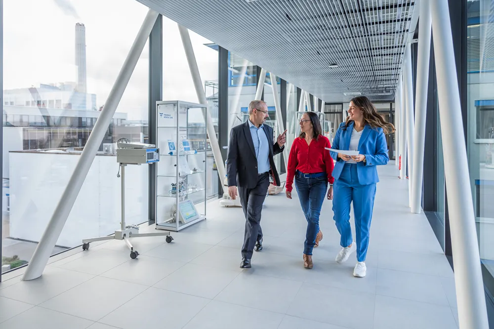 Three people in business attire walk and talk in a bright, modern hallway with large windows and display cases, suggesting a professional or corporate environment.