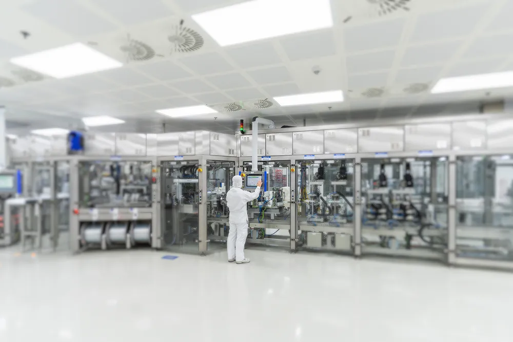 A person in full white protective clothing operates a touchscreen panel in a clean, modern laboratory or manufacturing facility with advanced automated machinery.