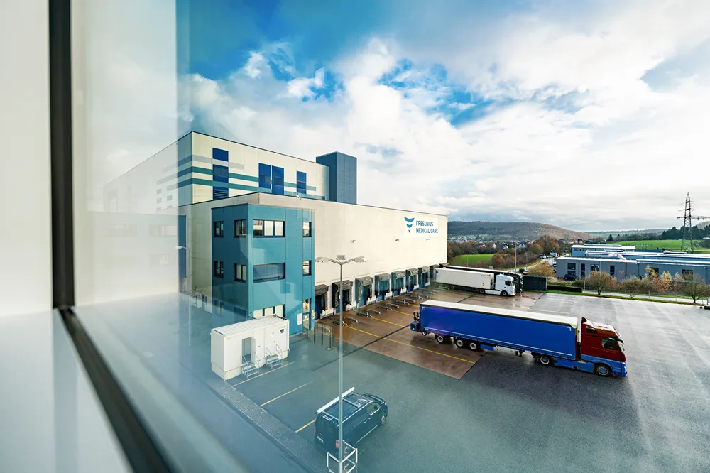 A modern industrial building labeled 'Fresenius Medical Centre' with loading docks, parked trucks, and a clear blue sky in the background. The photo is taken from behind a window, creating a slight reflection on the left.