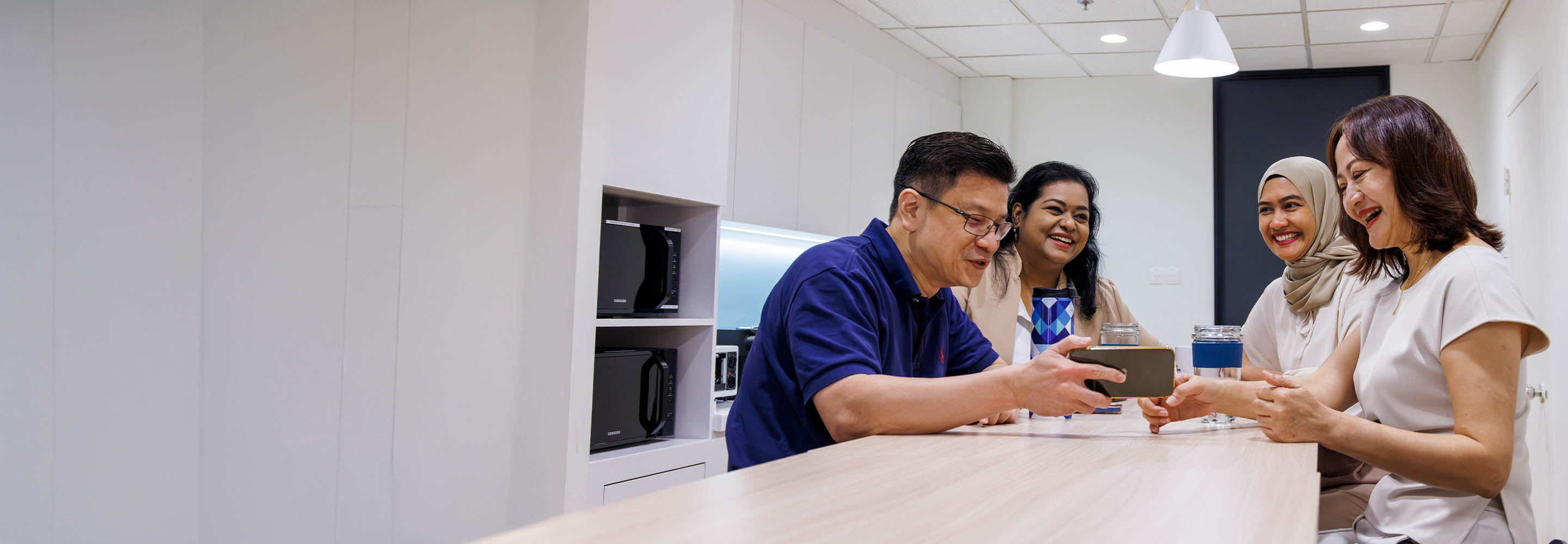 Four people sit around a table in an office break room, smiling and looking at a smartphone together. Two women wear headscarves, and everyone appears relaxed and happy.